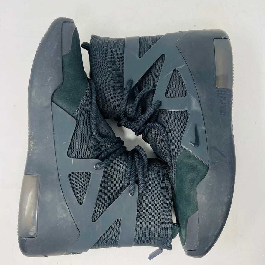 The Nike Air Fear of God 1 Triple Black is a high-top, dark, minimalist sneaker with angular overlays and a textured sole. It features a high collar, matte finish, dark laces, and has a small logo near the midsole. The background is light gray with general wear signs.