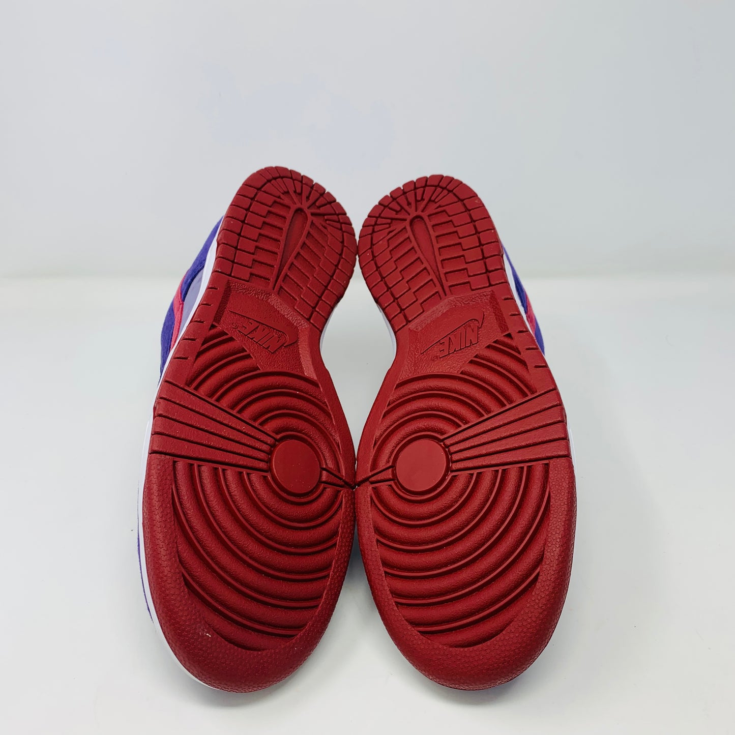 A pair of Nike Dunk Low Plum sneakers, in purple and maroon, rests on a red shoe box featuring a white Nike logo on a plain background. The shoes showcase a red swoosh and white midsoles, confirming their authenticity from Nike.