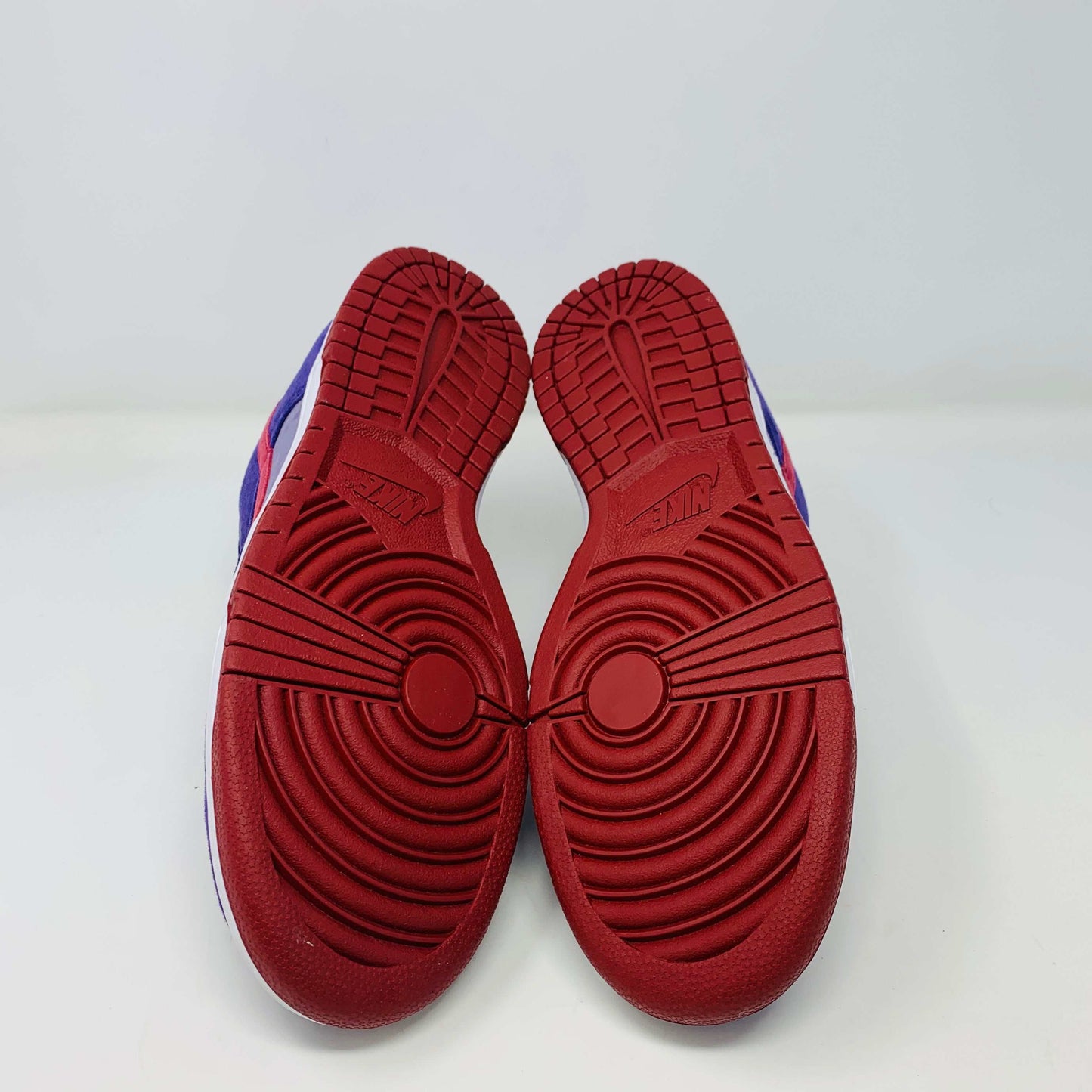 A pair of Nike Dunk Low Plum sneakers, in purple and maroon, rests on a red shoe box featuring a white Nike logo on a plain background. The shoes showcase a red swoosh and white midsoles, confirming their authenticity from Nike.