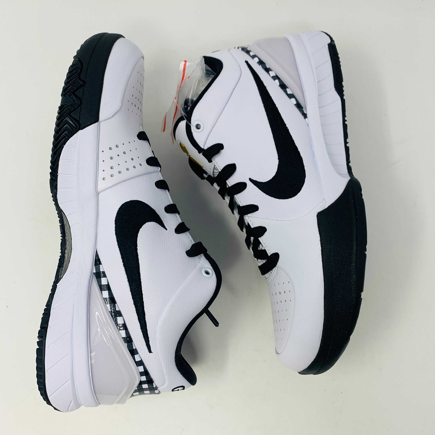 The Nike Kobe 4 Protro Mambacita Gigi shoe, featuring a black swoosh, black accents, and a checkered heel pattern, is displayed on a white box with a gold Nike logo. The box also includes extra laces.