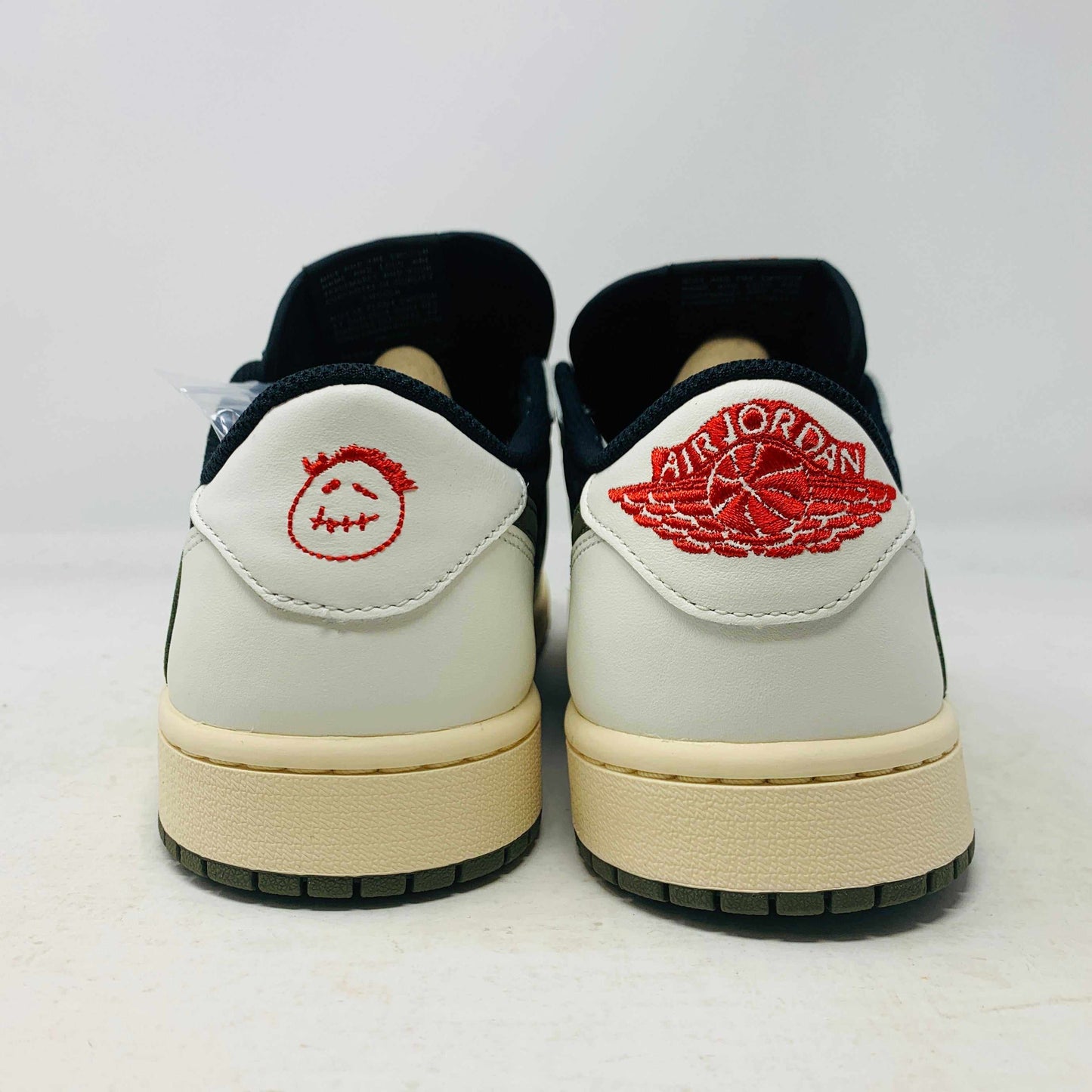 A black, white, and olive green Jordan 1 Retro Low OG SP Travis Scott Olive (W) sneaker from Nike is displayed on a matching box. This brand new shoe sports a large reversed swoosh and cream-colored sole. The box showcases the green Nike logo with 1985 Air Jordan 1 text for these authentic 2023 shoes.