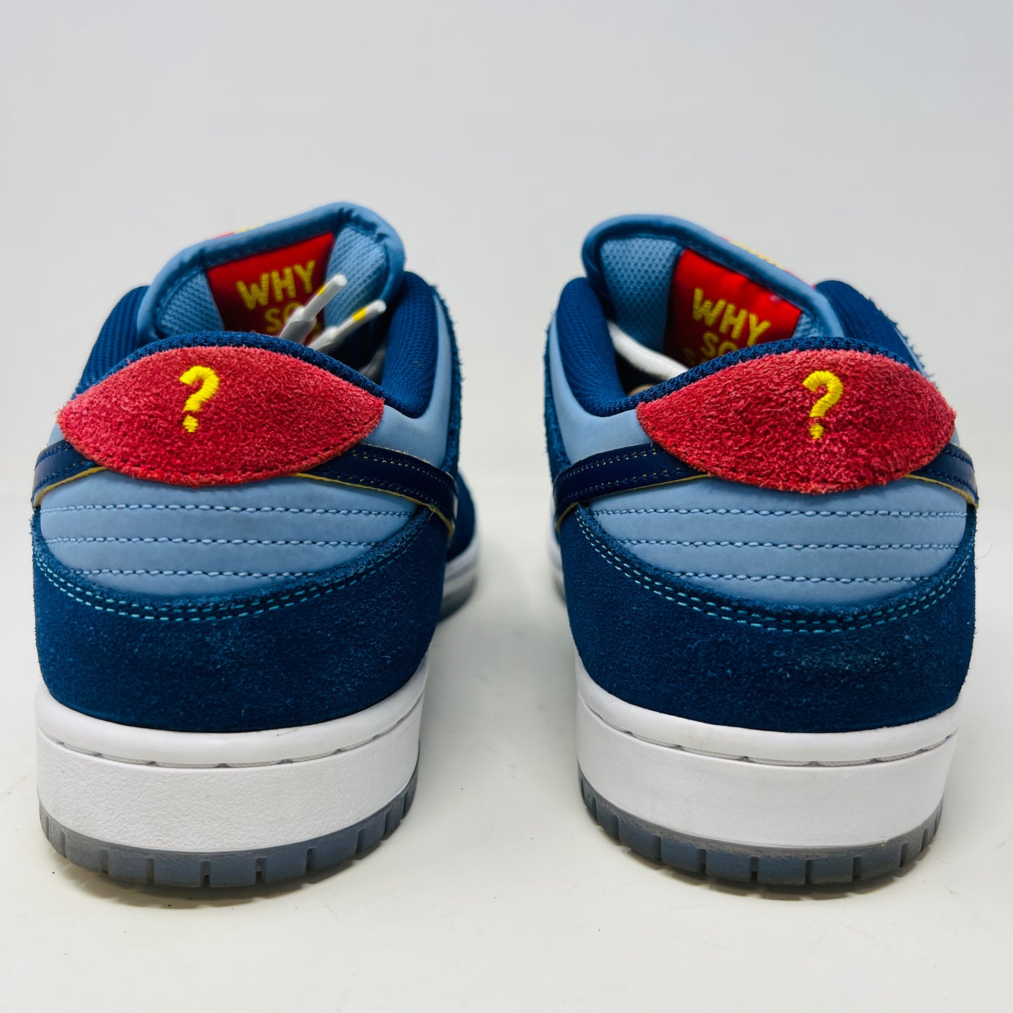 A Nike SB Dunk Low Pro Why So Sad? shoe, with blue uppers and white sole, rests on a purple box. It features an embroidered graphic on the heel, a dark blue swoosh, and extra laces in front. Price tags from 2022 are visible beside it.