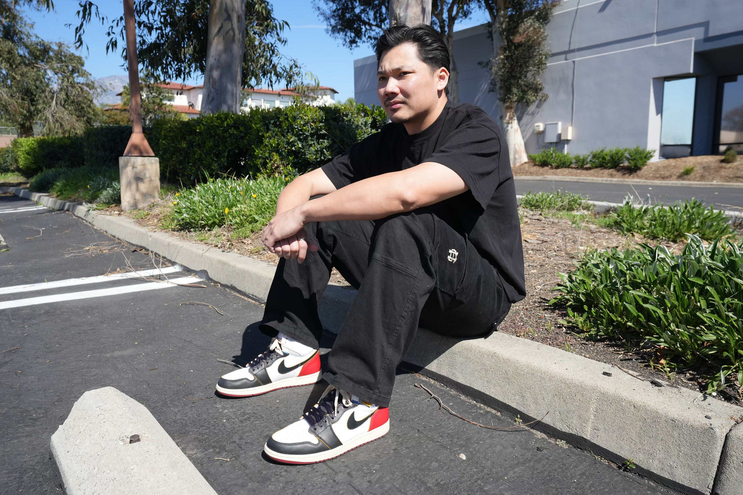 A person wearing a black outfit and sneakers with red, white, and black details sits on a curb in a parking lot. Theyre looking away, surrounded by greenery and buildings in the background.
