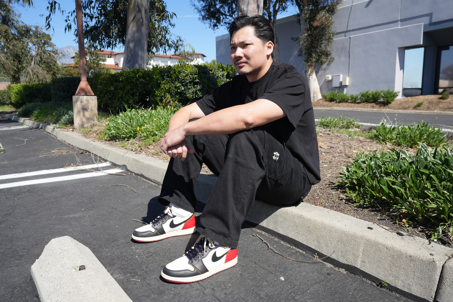 A person wearing a black outfit and sneakers with red, white, and black details sits on a curb in a parking lot. Theyre looking away, surrounded by greenery and buildings in the background.