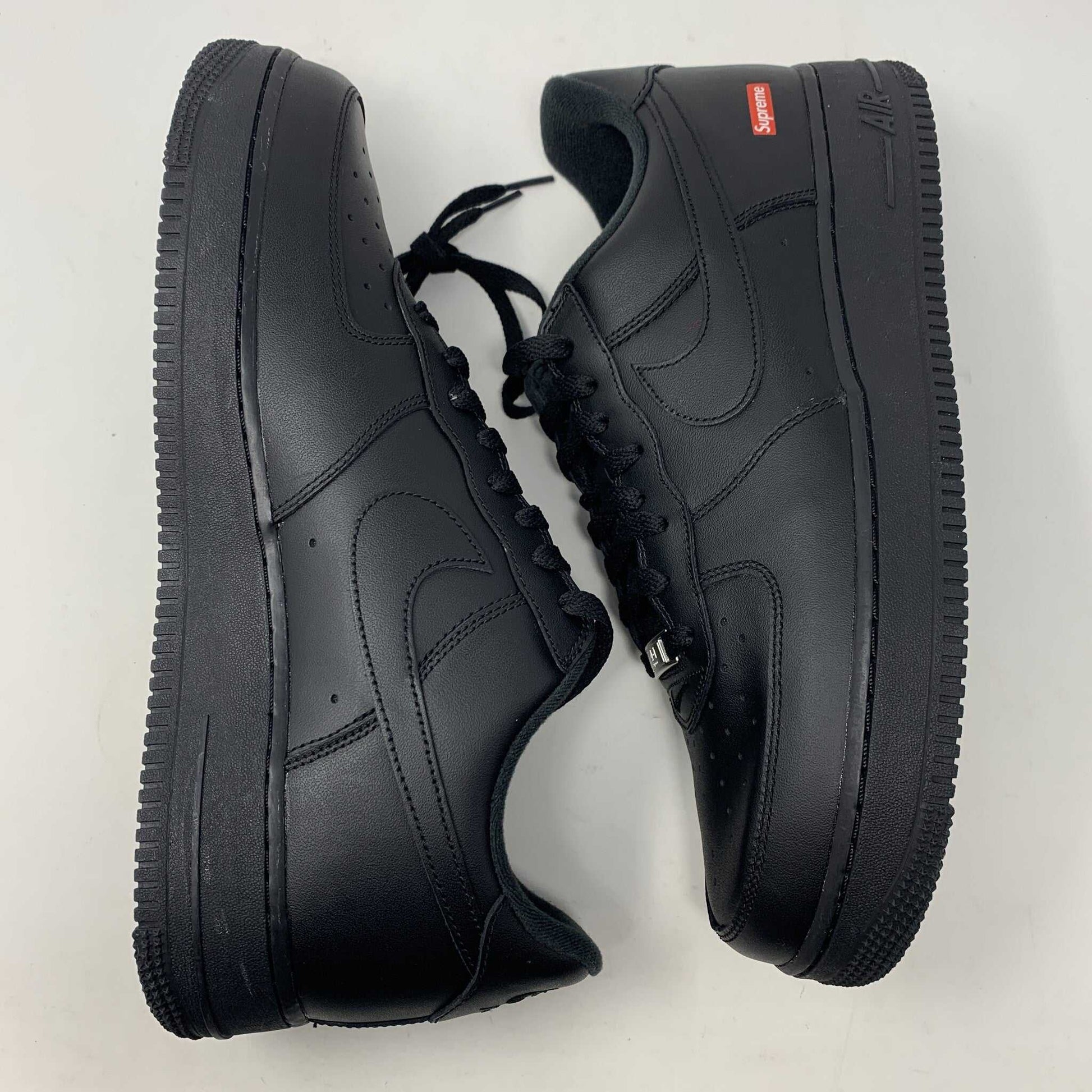 The Nike Supreme Air Force 1 Black features a perforated design on the toe box, black supreme laces with a shiny tag, and a prominent Nike logo, all set against a plain white background.