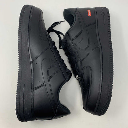 The Nike Supreme Air Force 1 Black features a perforated design on the toe box, black supreme laces with a shiny tag, and a prominent Nike logo, all set against a plain white background.