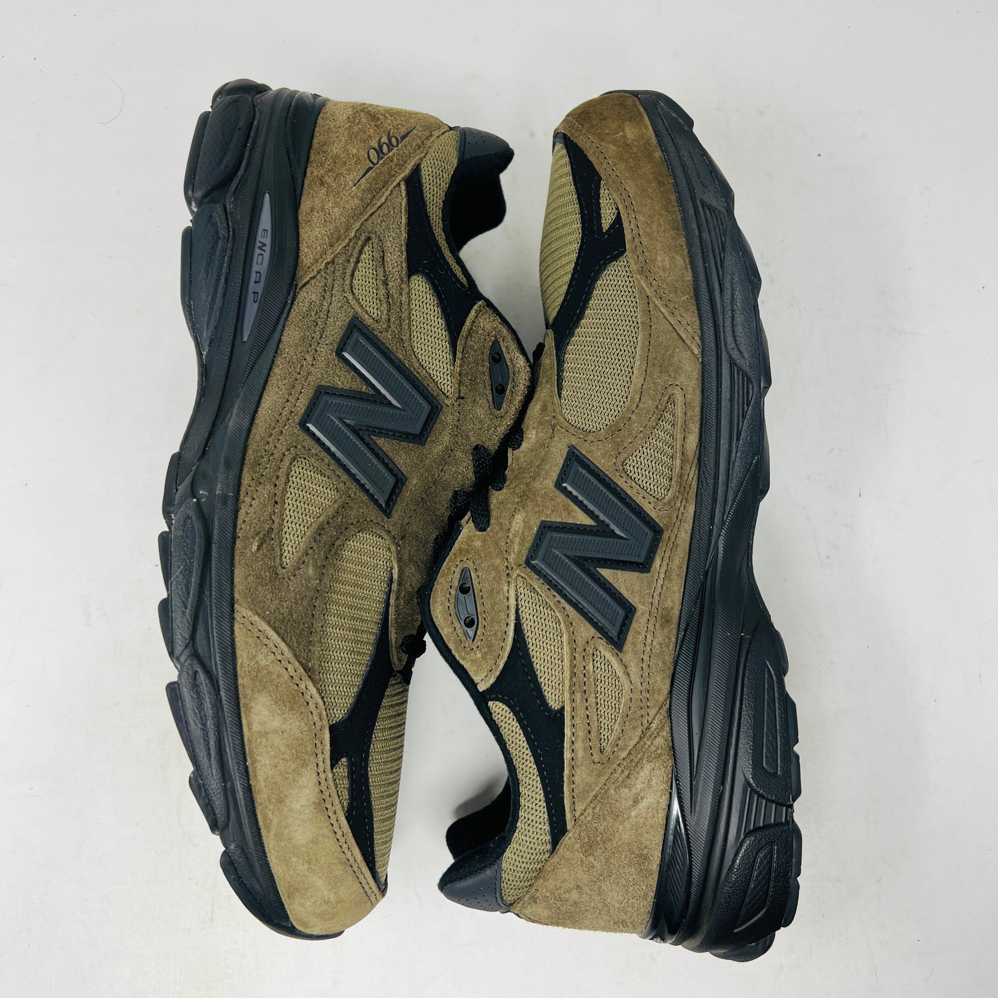 The New Balance 990v3 MiUSA JJJJound Brown Black athletic shoe, released in 2022, features tan and black mesh-suede uppers with an N logo on the side and 990 on the heel. It sits atop a black shoebox with the label M990JJ3 and shows light wear.