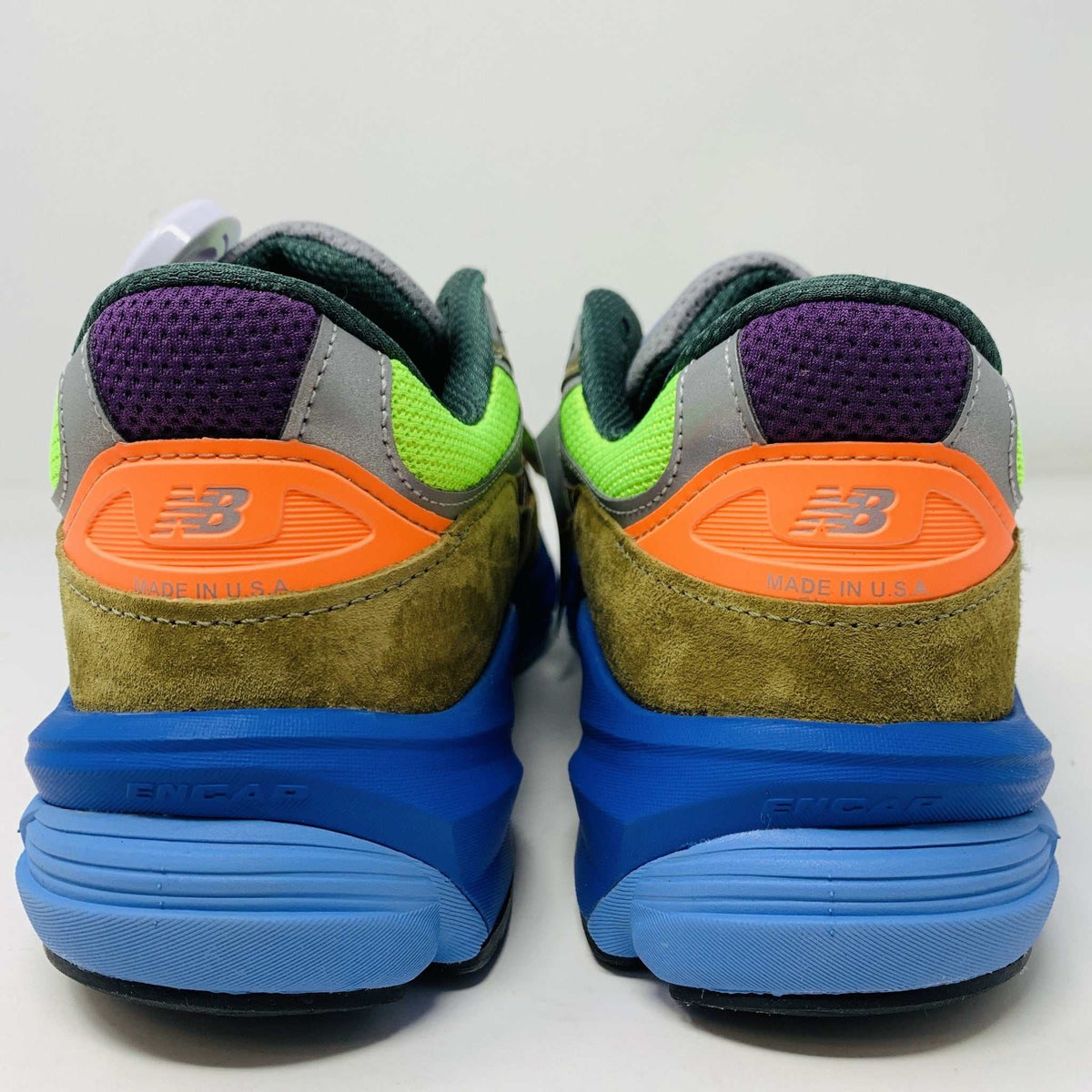 The New Balance 990v6 MiUSA Action Bronson Baklava sneaker, displayed on a white shoe box, boasts vibrant neon green, blue, and brown colors with suede and mesh materials. It features a distinctive design with a bold sole and branding on the side—truly authentic for 2023.