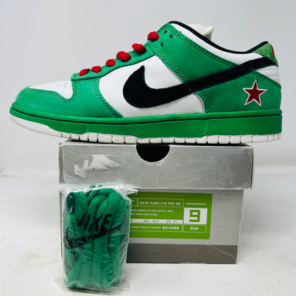 The Nike SB Dunk Low Heineken sneaker, featuring a green, white, and black design with red laces and a red star on the heel, sits on its box. A small package of green shoelaces is beside it. The box displays the size and Nike branding.