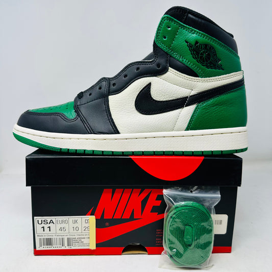 A pair of black, green, and white Jordan 1 Retro High Pine Green sneakers rests on a sleek black Nike shoebox. Featuring a prominent black swoosh and extra green laces, they have a faint smoke smell. The box lists size: USA 11, Euro 45, UK 10, CM 29.