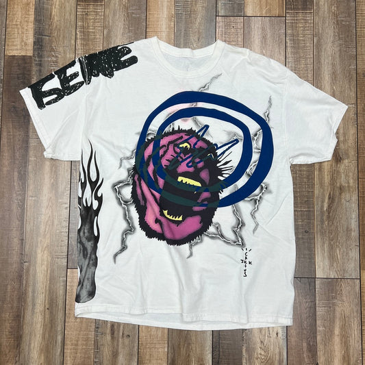 A preowned Travis Scott Cactus Jack For Fragment Sunrise Tee White is on a wooden floor, featuring a graphic of a screaming monster face with blue circles. Black flames line the sides, and ISENSE is boldly printed on the shoulder, capturing an edgy design vibe.