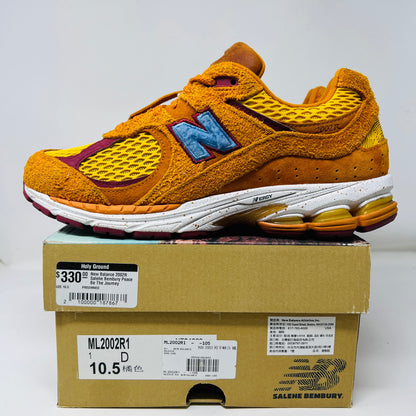 A New Balance 2002R Salehe Bembury Peace Be The Journey sneaker in vibrant orange and yellow with a blue N logo is displayed on a shoebox. It features textured design, visible sole cushioning, and extra laces. Tagged as model ML2002R1, size 10.5, priced at $330.