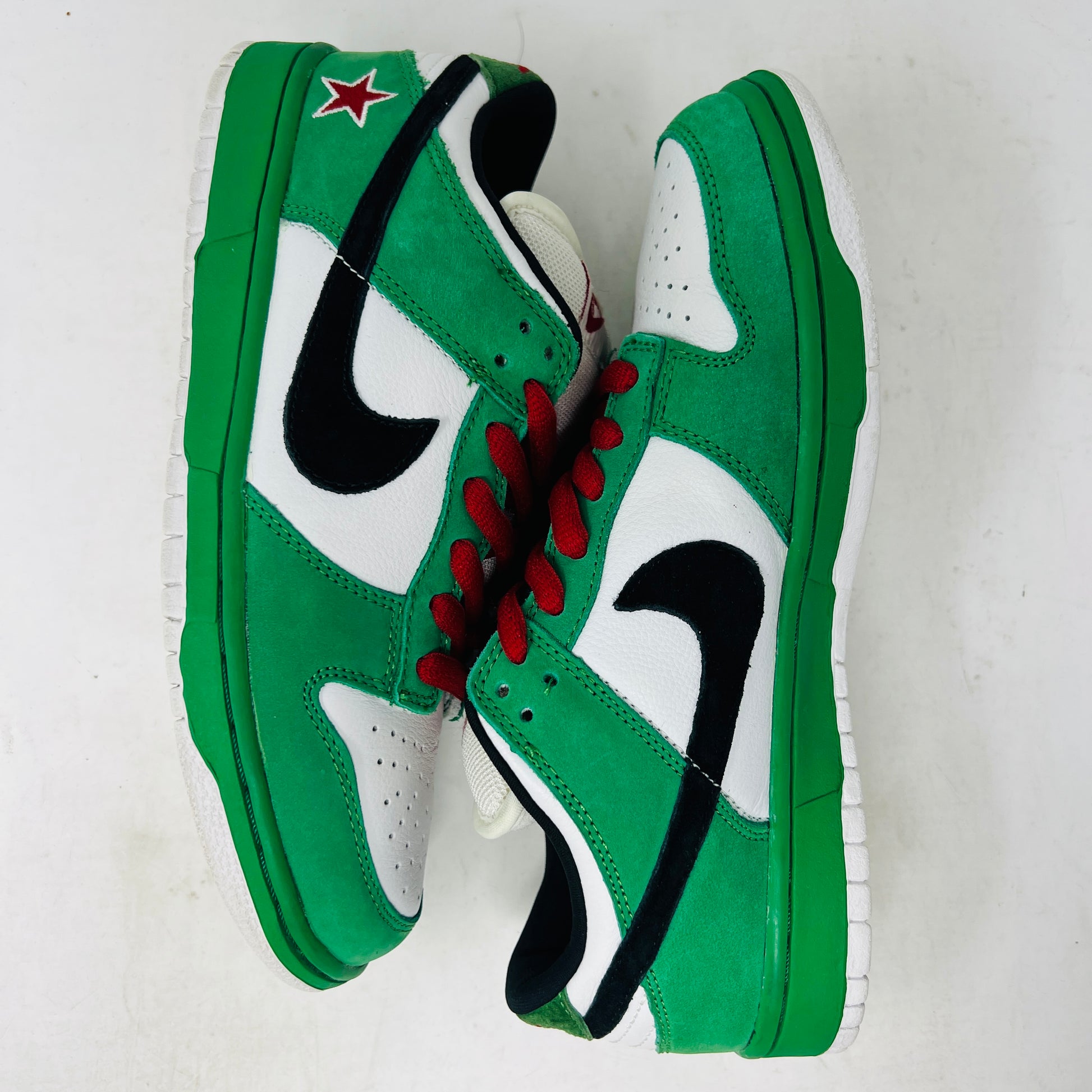 The Nike SB Dunk Low Heineken sneaker, featuring a green, white, and black design with red laces and a red star on the heel, sits on its box. A small package of green shoelaces is beside it. The box displays the size and Nike branding.