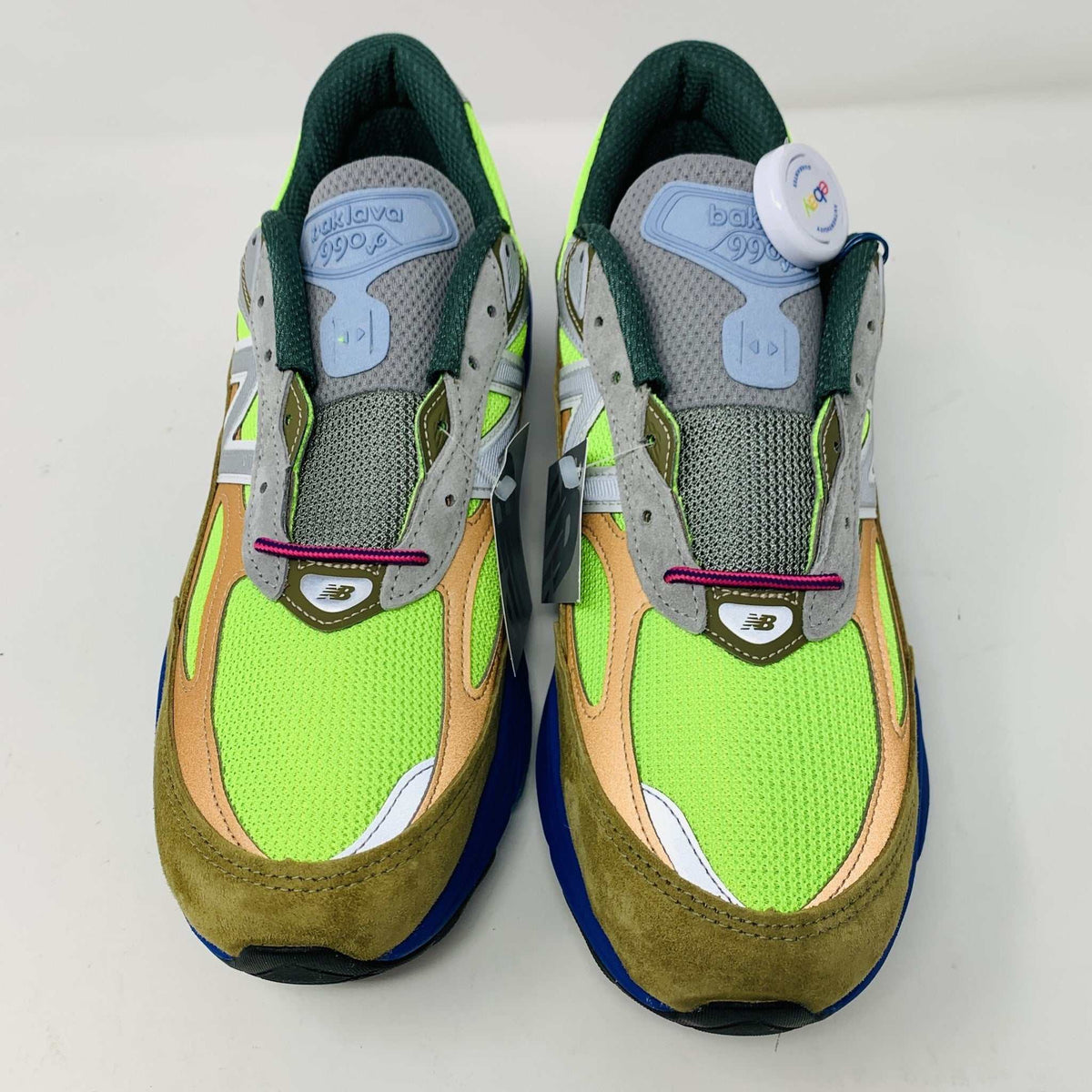 The New Balance 990v6 MiUSA Action Bronson Baklava sneaker, displayed on a white shoe box, boasts vibrant neon green, blue, and brown colors with suede and mesh materials. It features a distinctive design with a bold sole and branding on the side—truly authentic for 2023.