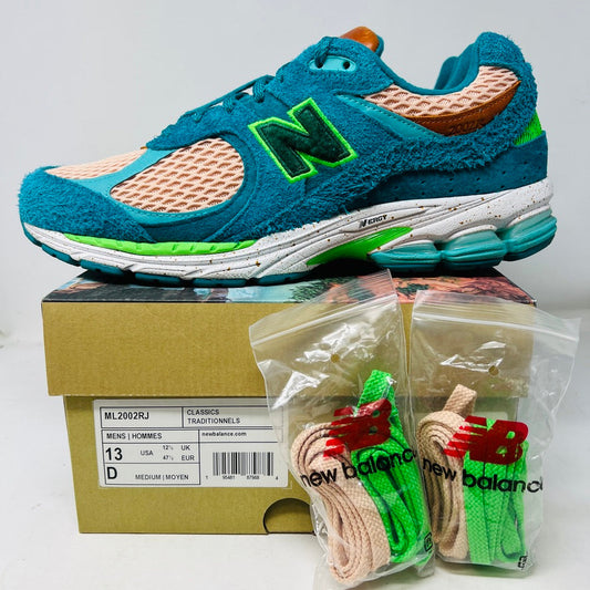 A pair of New Balance 2002R by Salehe Bembury, in colorful teal, pink, and green accents, sits atop a shoebox. Two extra lace sets—pink and green—are in front. The shoebox label details the size and style; the box is in good condition with a clean design.