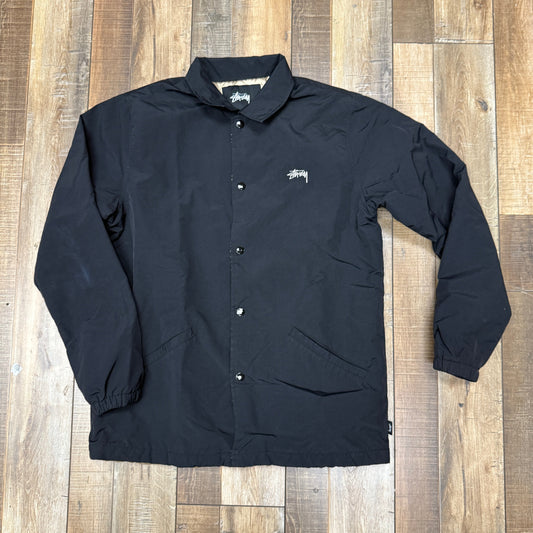 Black preowned Stussy Coaches Jacket on wooden floor.