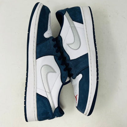 The Jordan 1 Low SB Midnight Navy sneakers, with white and navy panels and a side Nike swoosh, are displayed on a pristine shoebox. A spare set of white laces featuring the Nike SB logo in Metallic Silver is also included.