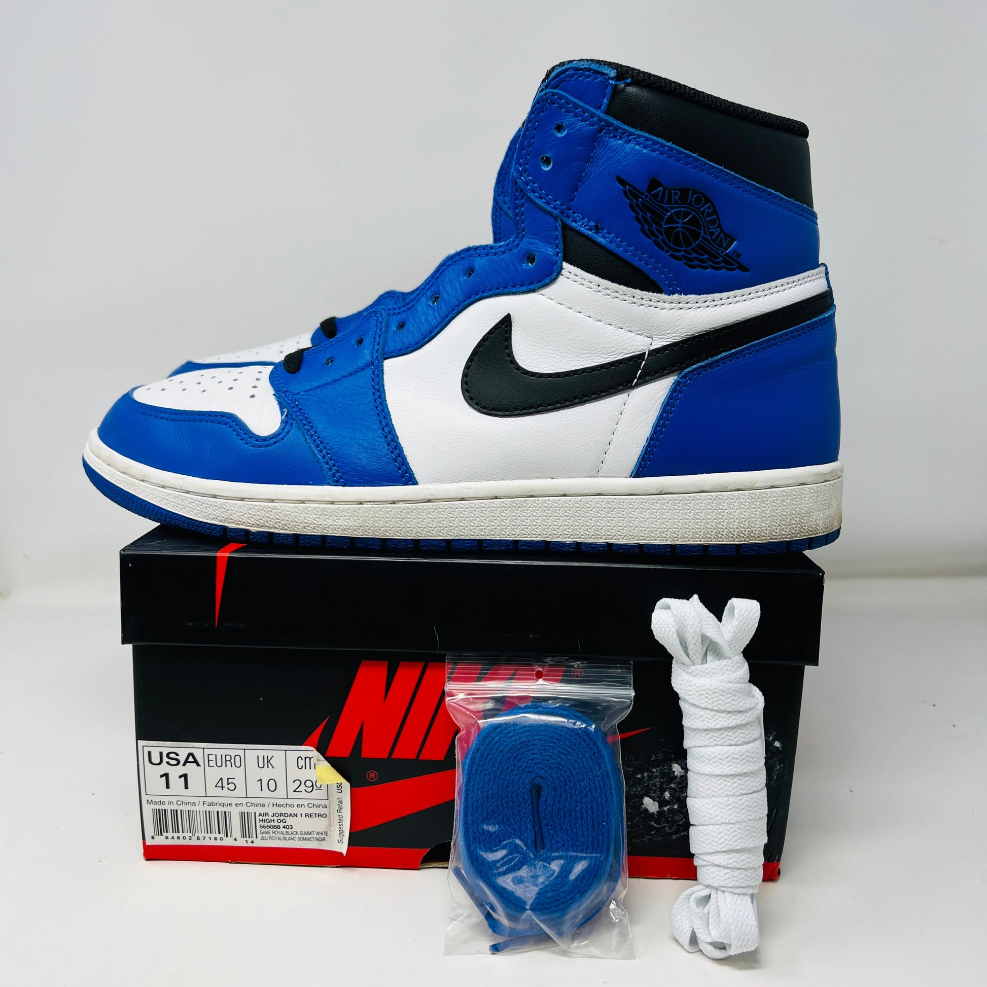 The Jordan 1 Retro High Game Royal high-top sneakers, in blue, white, and black with a clean upper, sit on a black and red box. Blue extra laces are bagged and white extra laces are laid out in front of the box, where size details are visible. Perfect for general wear.