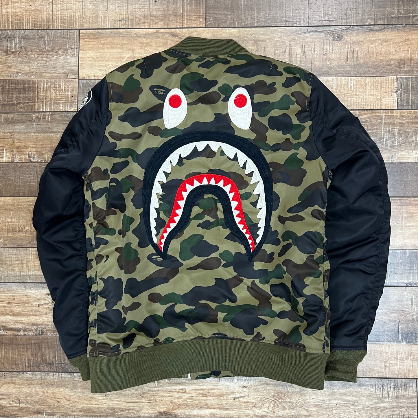 The preowned BAPE Shark MA-1 Jacket by Bape in green and brown camo features a black sleeve with the embroidered WGM letters and a cartoon tiger design, all displayed on a wooden floor.