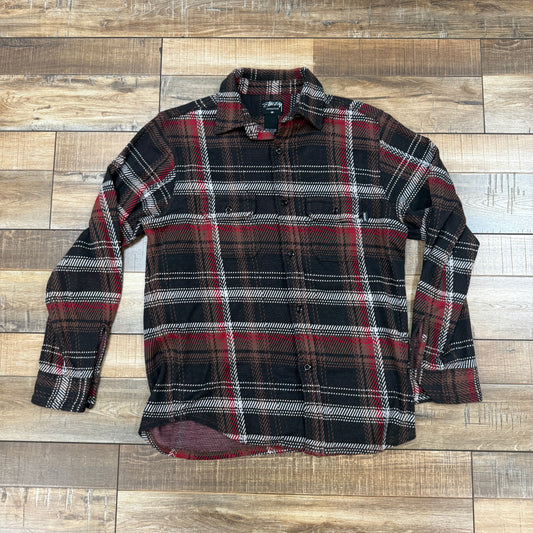 The Stussy Flannel Black/Red is a preowned long-sleeved shirt showcasing a classic blend of black, red, and white plaid, displayed flat on a wooden floor.