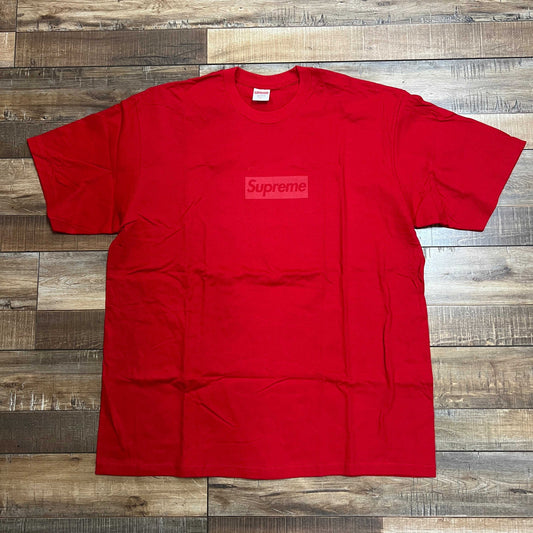 A pristine Supreme Tonal Box Logo Tee in red, with a subtle logo, lies flat and slightly wrinkled on a rustic wooden floor. Its bold hue beautifully contrasts against the varying brown shades of the textured surface.