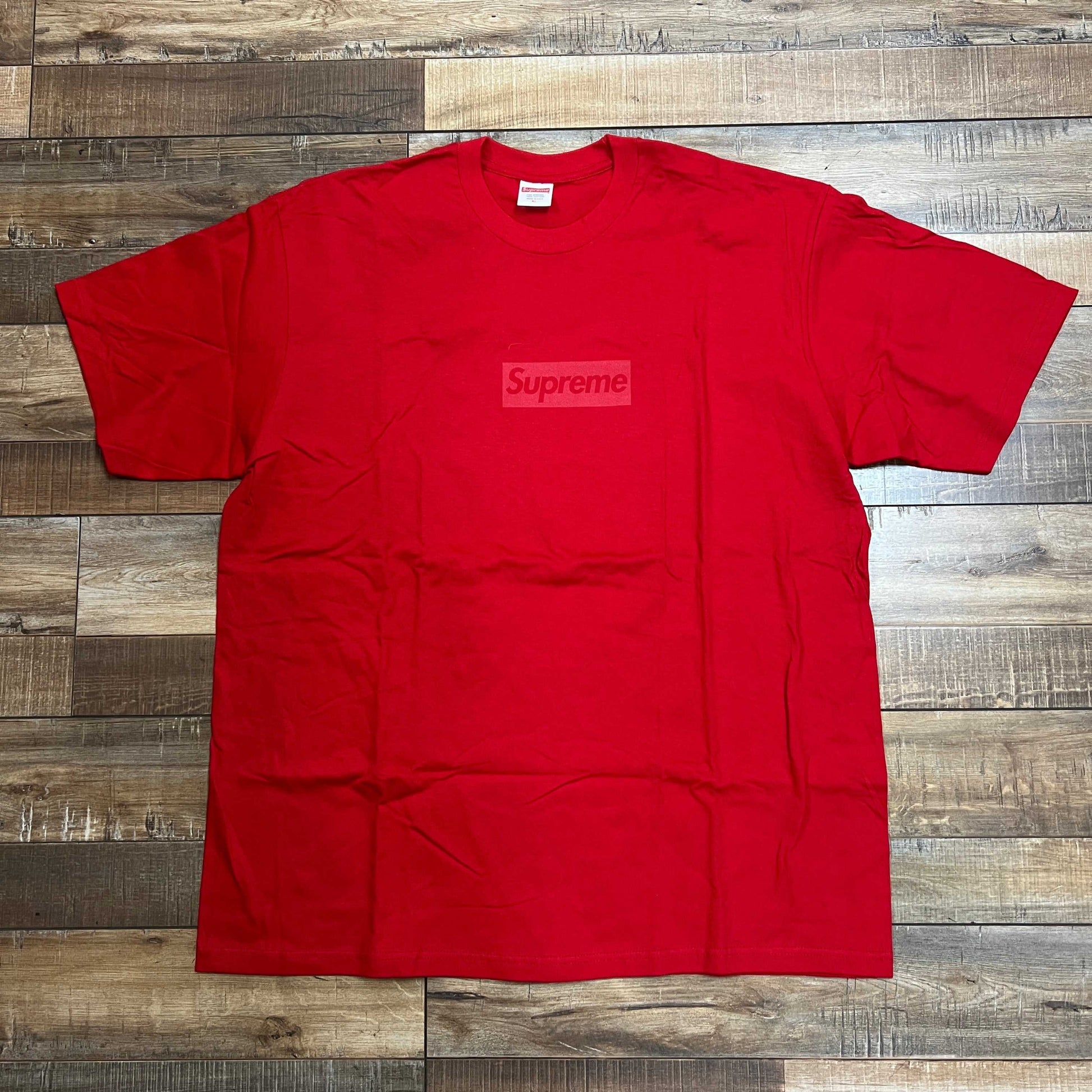 A pristine Supreme Tonal Box Logo Tee in red, with a subtle logo, lies flat and slightly wrinkled on a rustic wooden floor. Its bold hue beautifully contrasts against the varying brown shades of the textured surface.
