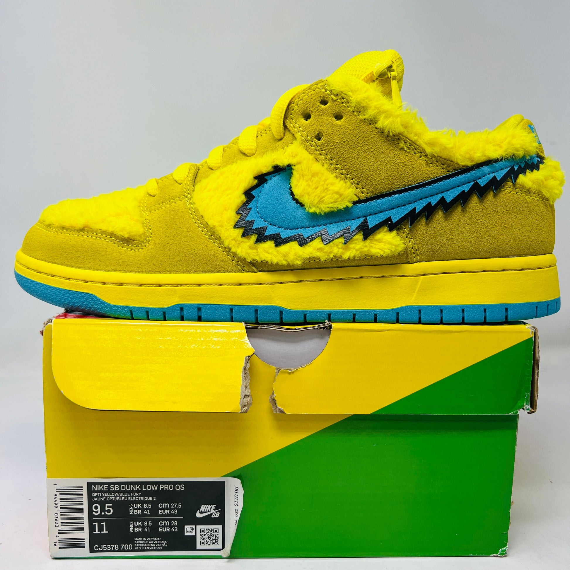 The Nike SB Dunk Low Grateful Dead Bears Opti Yellow sneaker shines brand new with fuzzy detailing and a blue swoosh atop its colorful but slightly damaged box. Released in 2020, this vibrant yellow shoe by Nike is partially visible in its packaging.