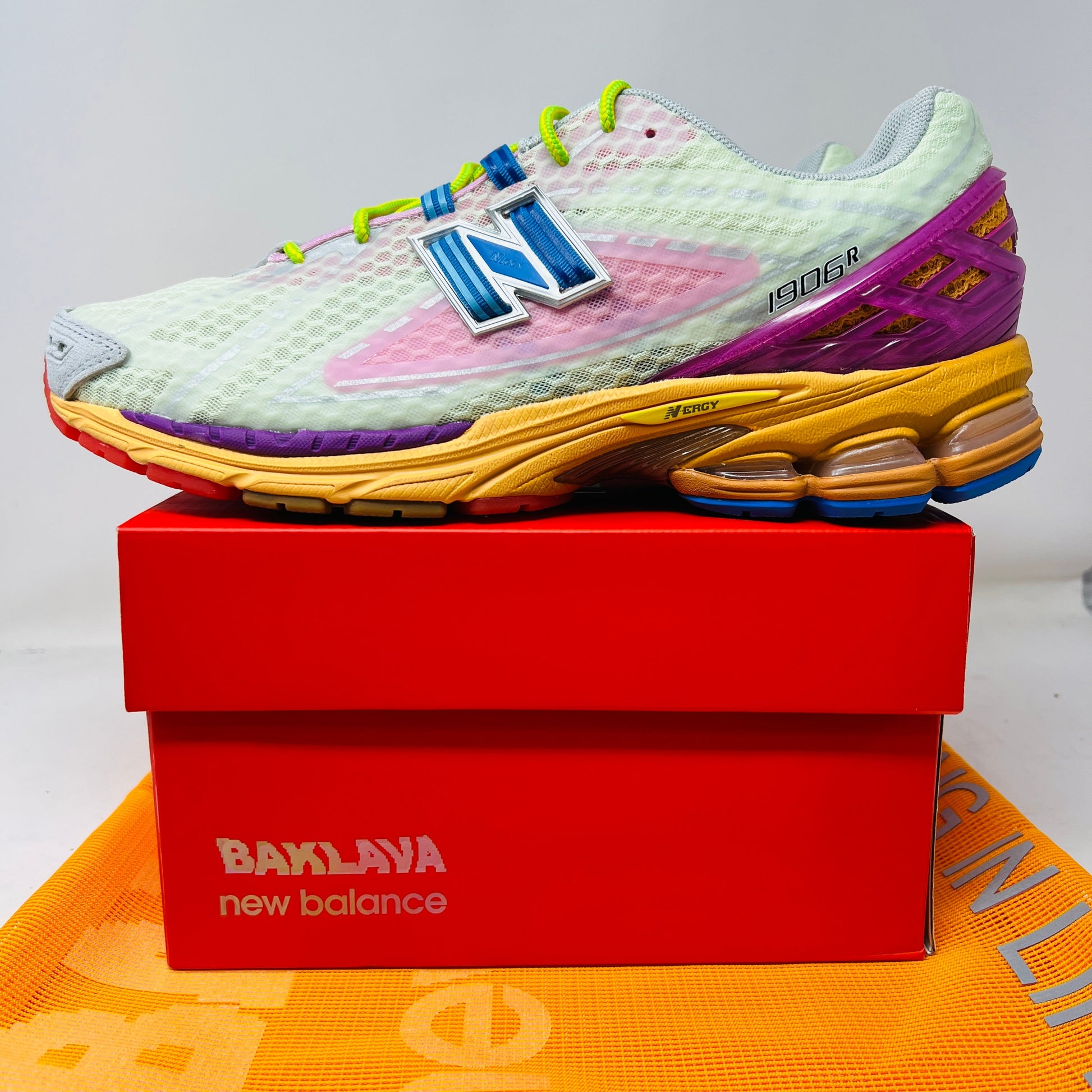 The New Balance 1906R Action Bronson Rosewater sneaker, boasting a light mesh upper and colorful sole with a bold blue N logo, rests on a pristine red Baylaya box. An orange surface partially peeks out under the box.