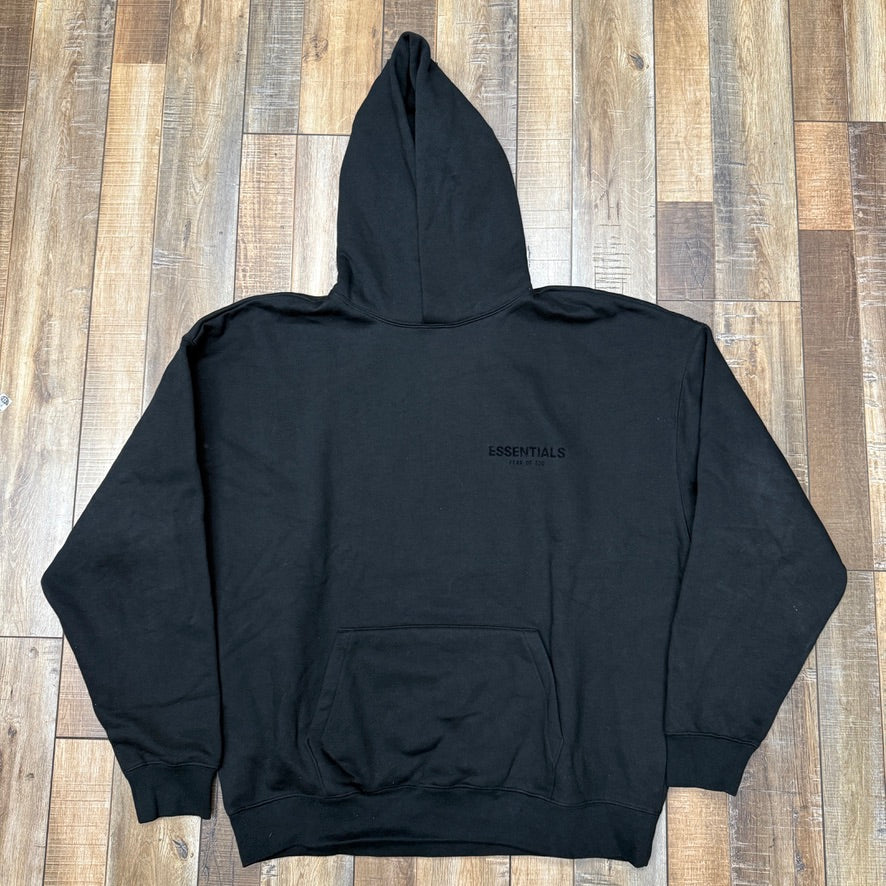 Fear of God Essentials Logo Hoodie in Stretch Limo/Black, laid flat on a wooden floor, featuring ESSENTIALS text on the back, a large front pocket, and a hood.