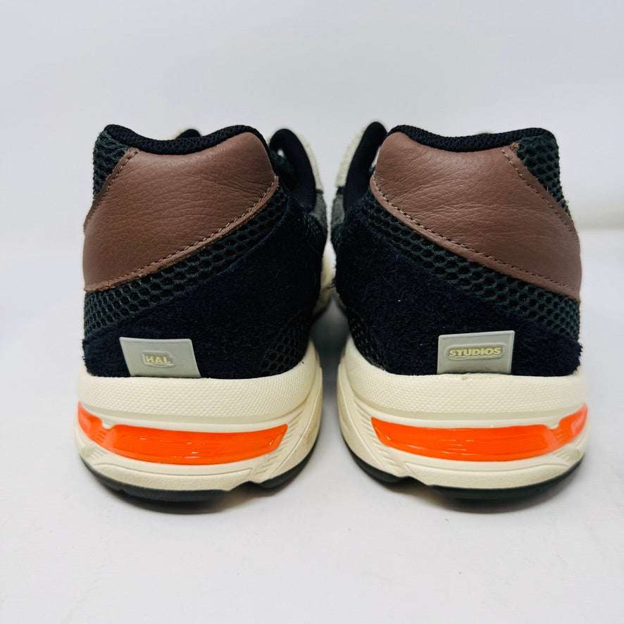 A sleek ASICS Gel-1130 MK-II HAL Studio Forest sits on its box, displaying black and brown mesh with suede accents. Distinctive orange detailing highlights the heel, while an extra brown laces bag accompanies it, enhancing its authentic athletic style.