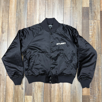 The Stussy Souvenir Jacket FW24 by Holy Ground Sneaker Shop is a black satin bomber with ribbed cuffs, collar, and hem. It features a snap button front and Stussy embroidered in white on the left chest. The jacket is laid flat on a wooden floor.