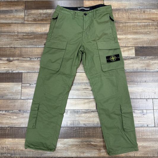 Brand new Stone Island Cargo Pants Green with multiple pockets are displayed on a wooden floor, featuring a compass patch on one leg, available at Holy Ground Sneaker Shop.