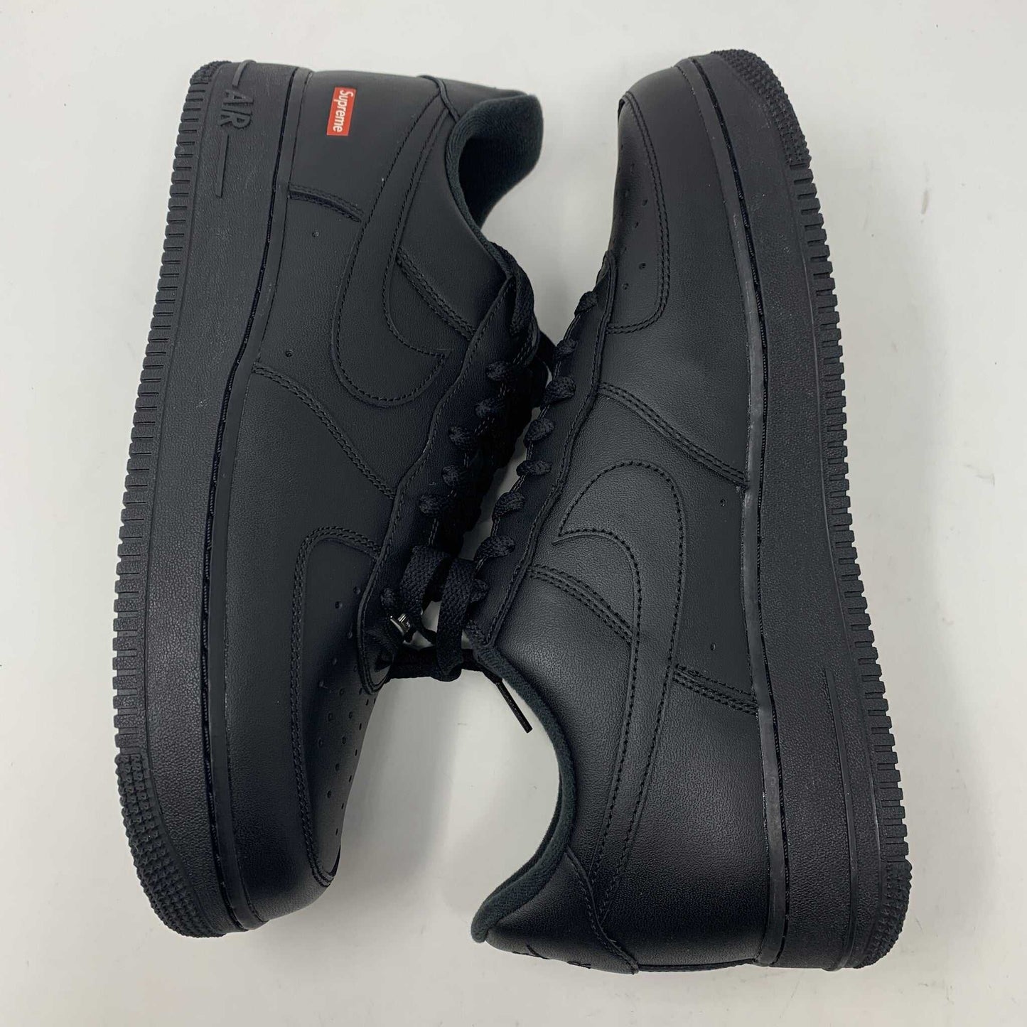 The Nike Supreme Air Force 1 Black features a perforated design on the toe box, black supreme laces with a shiny tag, and a prominent Nike logo, all set against a plain white background.