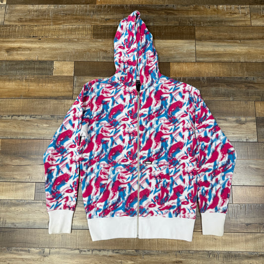 The Stussy Psychedelic All Over Hoodie, from the brand Stussy, showcases a vibrant abstract pattern in shades of pink, blue, and white. It features a zip-up front and hood and is displayed on a wooden floor.