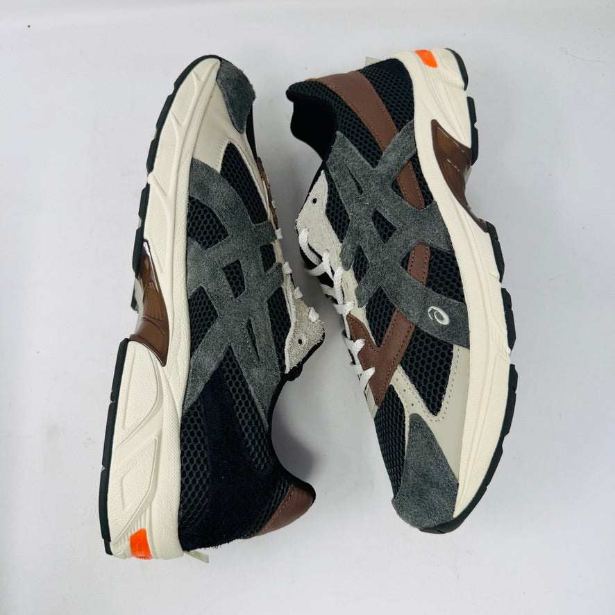 A sleek ASICS Gel-1130 MK-II HAL Studio Forest sits on its box, displaying black and brown mesh with suede accents. Distinctive orange detailing highlights the heel, while an extra brown laces bag accompanies it, enhancing its authentic athletic style.