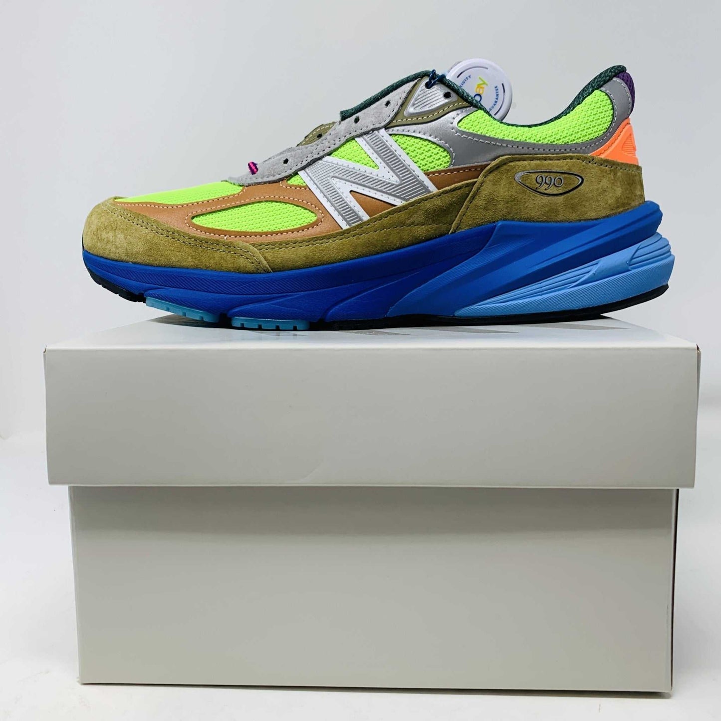 The New Balance 990v6 MiUSA Action Bronson Baklava sneaker, displayed on a white shoe box, boasts vibrant neon green, blue, and brown colors with suede and mesh materials. It features a distinctive design with a bold sole and branding on the side—truly authentic for 2023.
