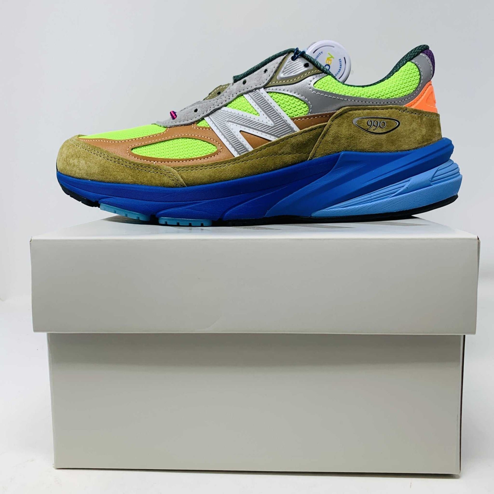 The New Balance 990v6 MiUSA Action Bronson Baklava sneaker, displayed on a white shoe box, boasts vibrant neon green, blue, and brown colors with suede and mesh materials. It features a distinctive design with a bold sole and branding on the side—truly authentic for 2023.