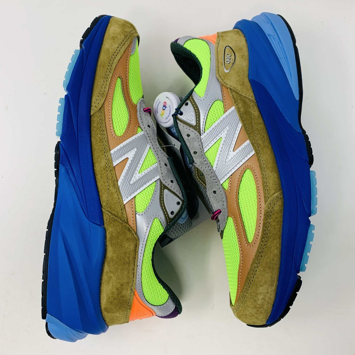 The New Balance 990v6 MiUSA Action Bronson Baklava sneaker, displayed on a white shoe box, boasts vibrant neon green, blue, and brown colors with suede and mesh materials. It features a distinctive design with a bold sole and branding on the side—truly authentic for 2023.
