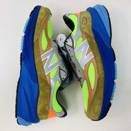 The New Balance 990v6 MiUSA Action Bronson Baklava sneaker, displayed on a white shoe box, boasts vibrant neon green, blue, and brown colors with suede and mesh materials. It features a distinctive design with a bold sole and branding on the side—truly authentic for 2023.