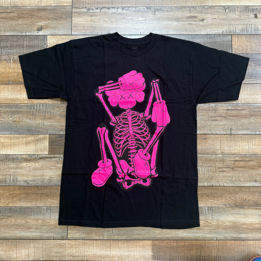 The KAWS SKELETON NEW FICTION T-shirt by Kaws is a black tee with a bright pink skeleton design, featuring cartoonish elements like X marks for eyes and exaggerated hands and feet, all showcased against a wooden floor.