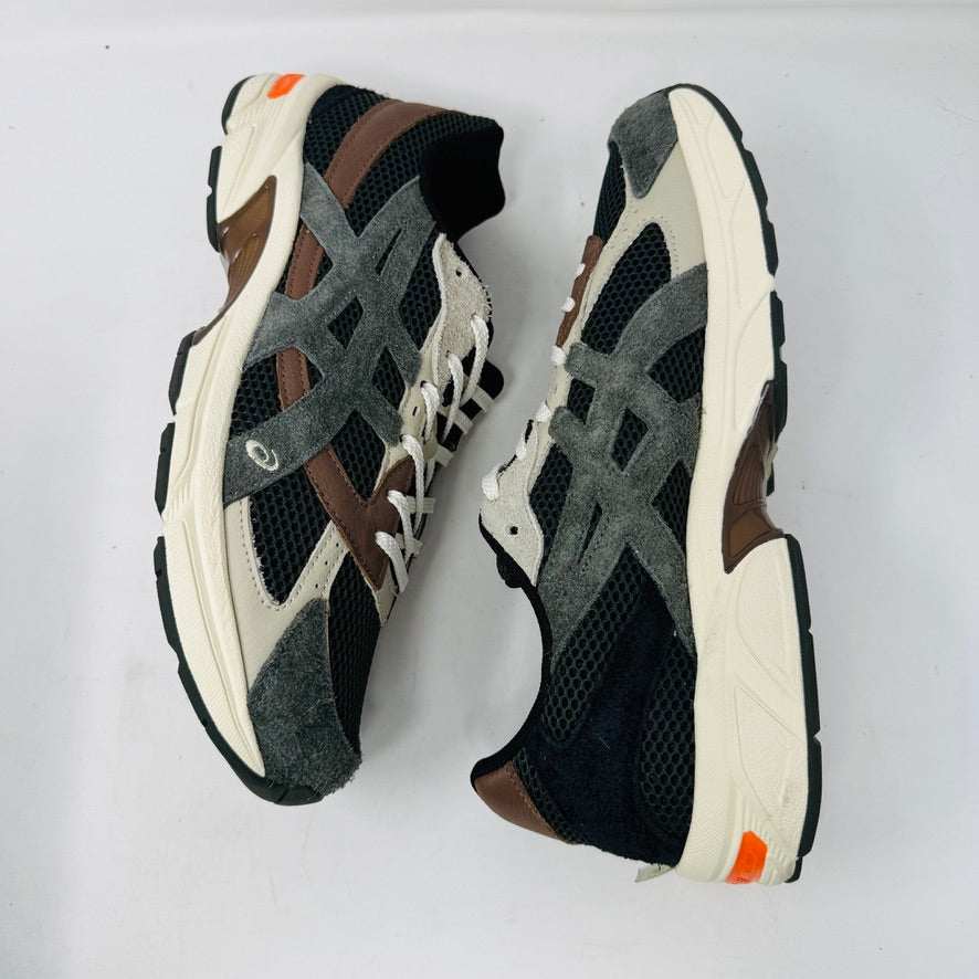 A sleek ASICS Gel-1130 MK-II HAL Studio Forest sits on its box, displaying black and brown mesh with suede accents. Distinctive orange detailing highlights the heel, while an extra brown laces bag accompanies it, enhancing its authentic athletic style.