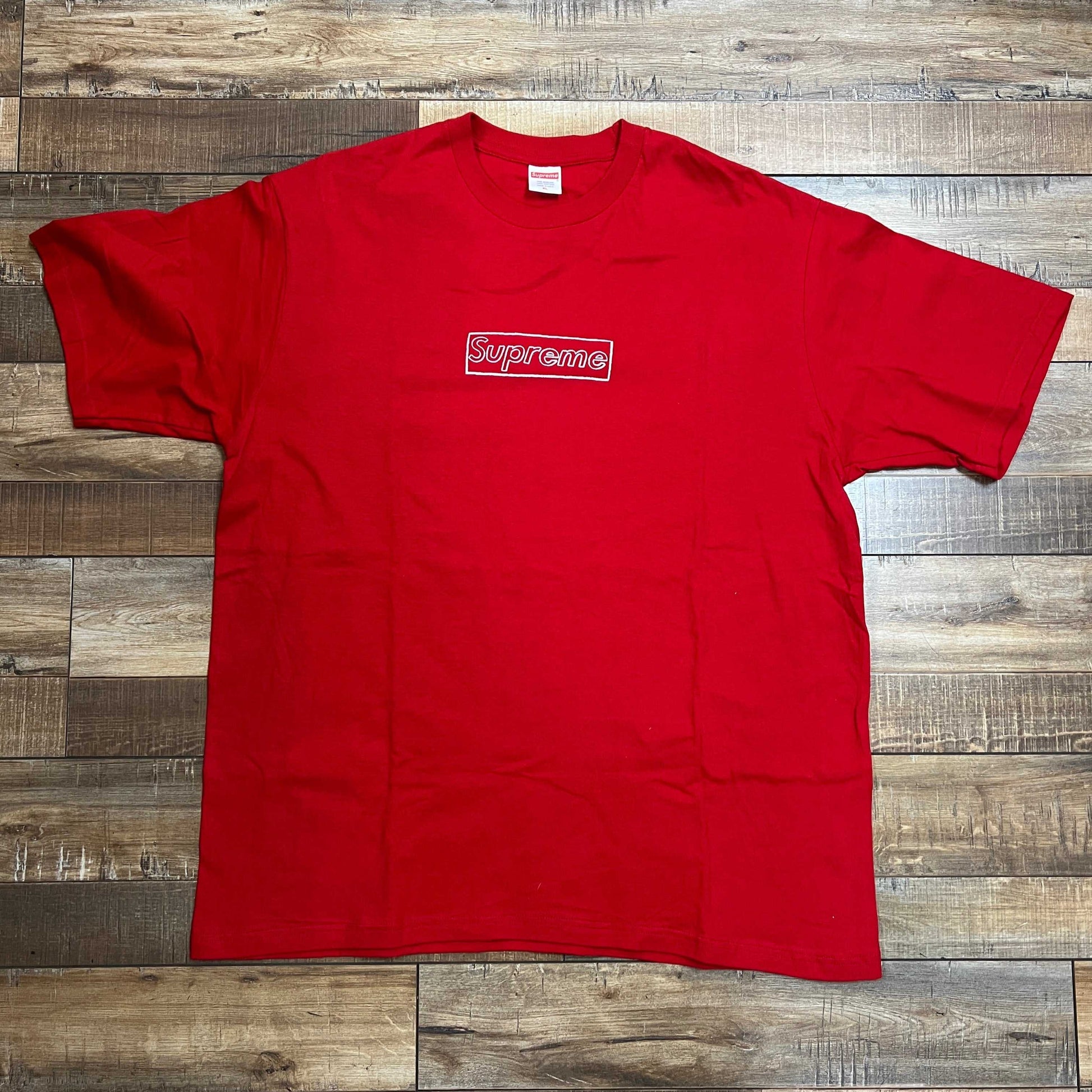 A Supreme KAWS Chalk Logo Tee in red, featuring the iconic logo in crisp white lettering inside a red box, is beautifully laid out on a wooden floor.
