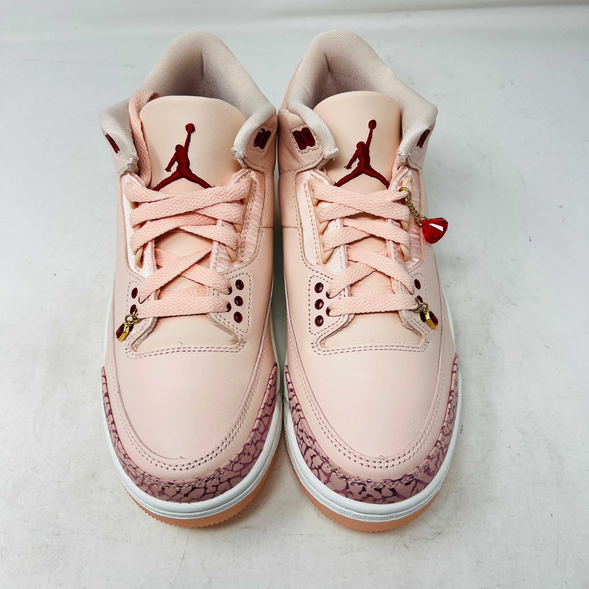 The BRAND NEW Jordan 3 Retro Valentines Day Treat Yourself (2025) (Womens) by Jordan, in pink and cream with textured heel and toe patterns, sits on a closed red shoebox. It features a translucent sole, red eyelets, and an authentic pendant on the laces sparkling like GEMS.