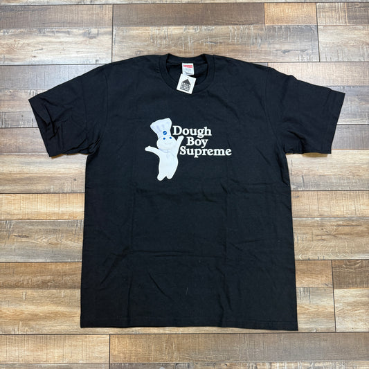 The Supreme Doughboy Tee Black features a printed doughboy wearing a chefs hat alongside Dough Boy Supreme text in white, laid flat on a wooden floor.