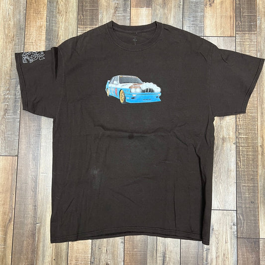 Preowned Travis Scott JACKBOYS Vehicle T-shirt in brown by Cactus Jack, featuring a vibrant car illustration with blue front and notable headlights. Laid flat on wood, it has a small white graphic design on the sleeve for added charm.