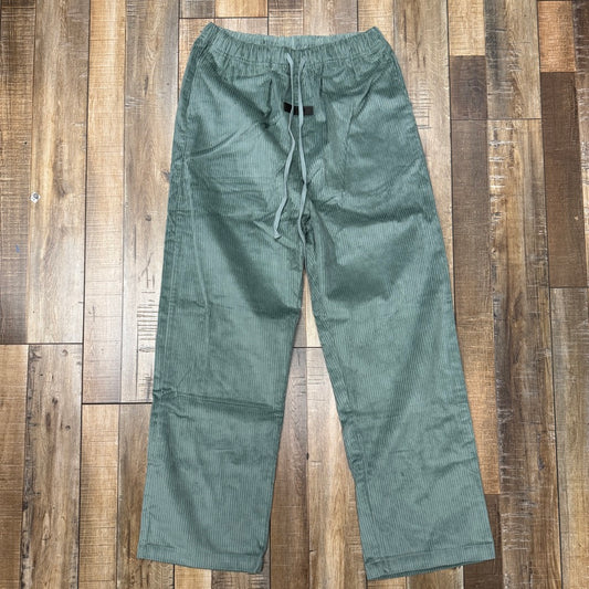 Laid flat on a wooden floor, the Essentials Fear of God Relaxed Trouser in Sycamore features a drawstring waist and straight-leg fit, complete with subtle horizontal stripes in the green corduroy fabric.