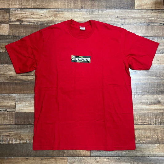 The Supreme Box Logo Tee (FW23) in red features a black box logo with Supreme in white text on the chest, displayed on a wooden floor.