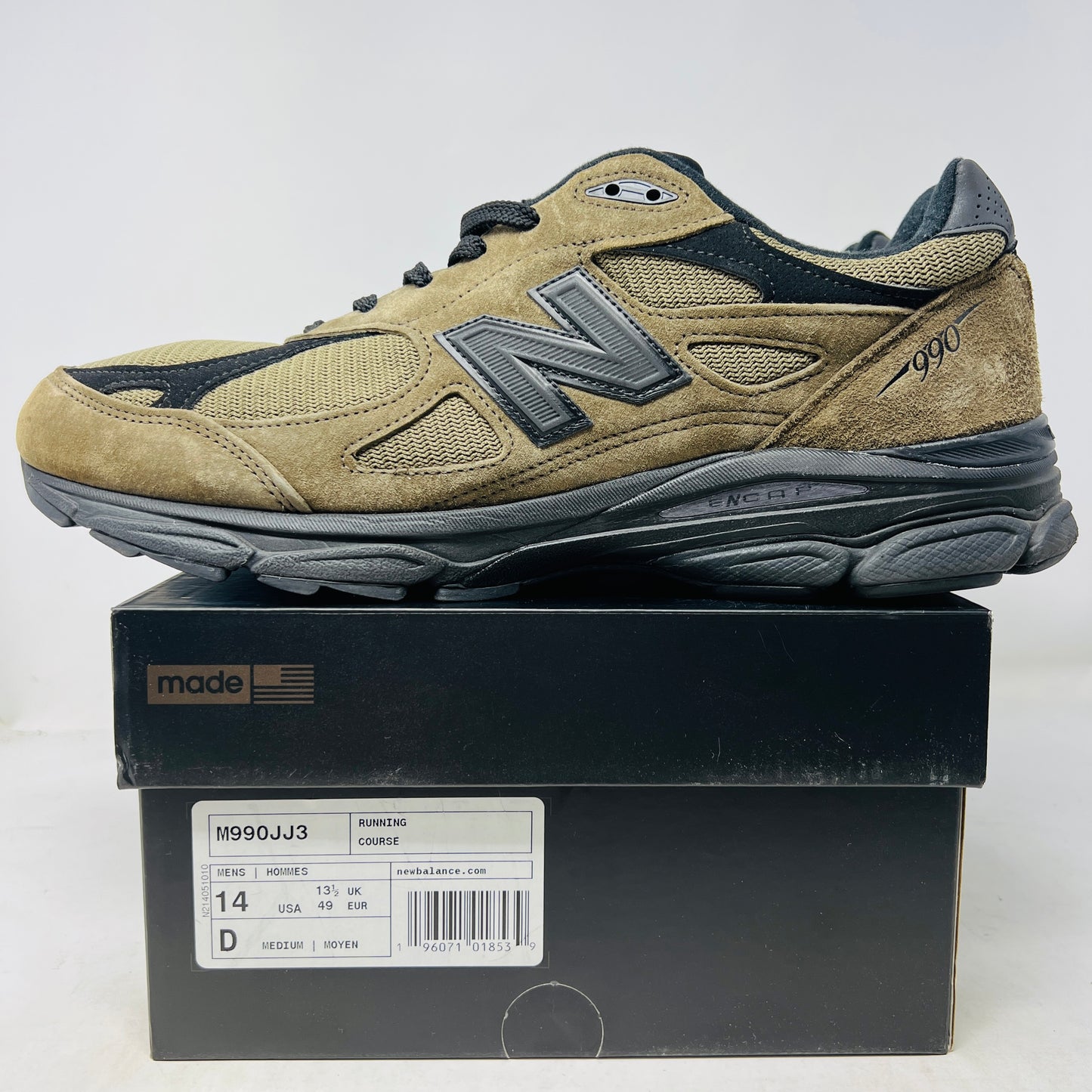 The New Balance 990v3 MiUSA JJJJound Brown Black athletic shoe, released in 2022, features tan and black mesh-suede uppers with an N logo on the side and 990 on the heel. It sits atop a black shoebox with the label M990JJ3 and shows light wear.
