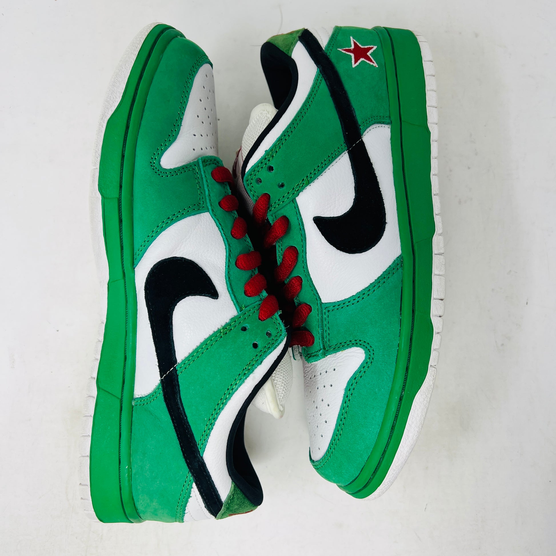 The Nike SB Dunk Low Heineken sneaker, featuring a green, white, and black design with red laces and a red star on the heel, sits on its box. A small package of green shoelaces is beside it. The box displays the size and Nike branding.