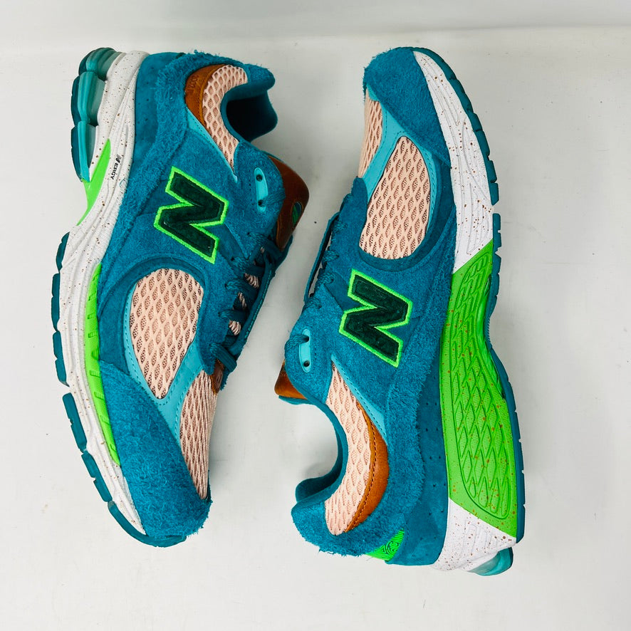 A pair of New Balance 2002R by Salehe Bembury, in colorful teal, pink, and green accents, sits atop a shoebox. Two extra lace sets—pink and green—are in front. The shoebox label details the size and style; the box is in good condition with a clean design.